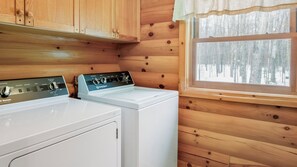 A laundry room with two white washing machines, wooden walls, and a window with a view of trees.