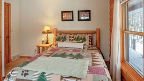 A cozy bedroom with a wooden headboard, a quilt with nature patterns, a bedside lamp, and two framed pictures on the wall.