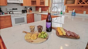 A kitchen counter with a bottle of wine, a cheese board, crackers, and a plate of grapes.