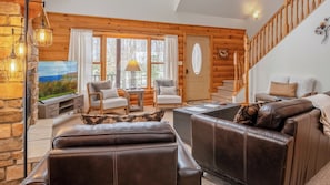 A cozy log cabin interior with a fireplace, leather sofas, a dining area, and a staircase.