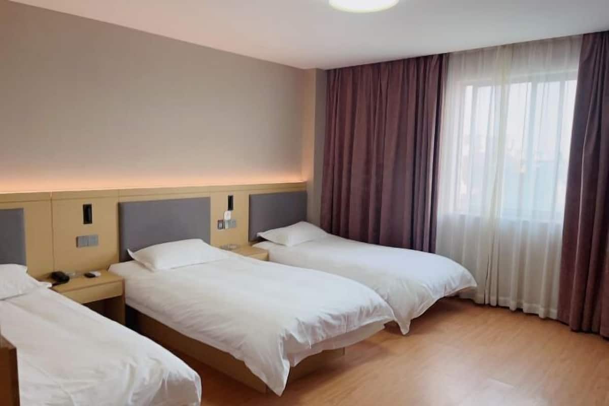Une chambre d’hôtel avec trois lits, un plancher en bois, une grande fenêtre avec des rideaux et une table de chevet avec un téléphone.