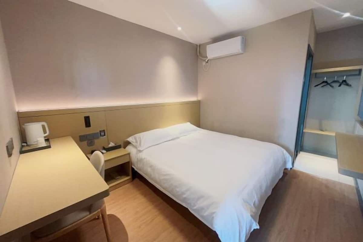 Une chambre d’hôtel comprenant un lit, un bureau, une chaise, une table de chevet, un climatiseur fixé au mur et un porte-manteau.