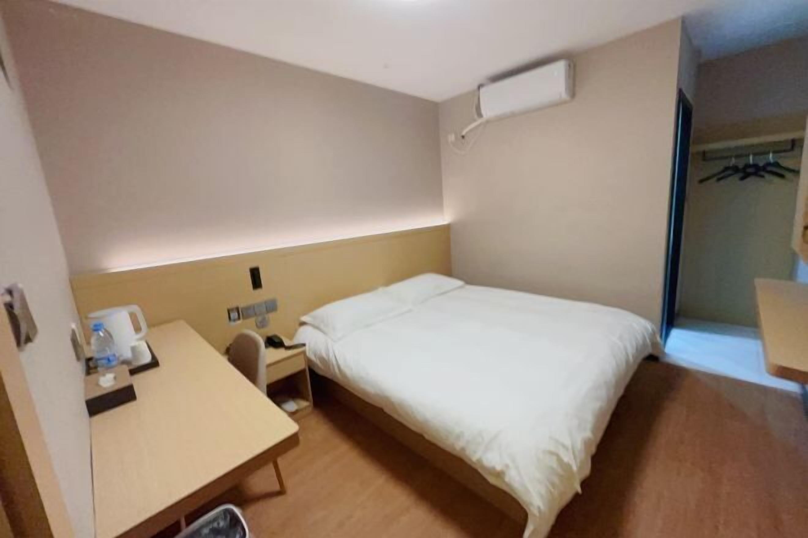 A hotel room with a bed, a desk, a chair, a trash bin, and a coat rack.