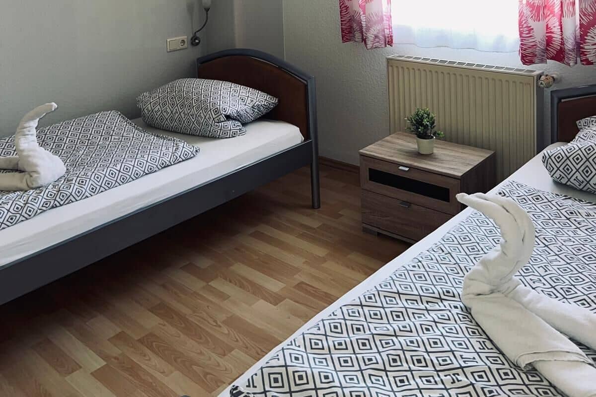 A bedroom with two single beds, a small bedside table with a plant, and a window with red curtains.