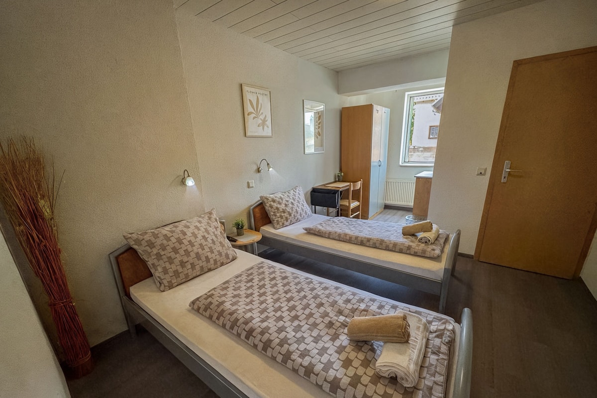 A room with two beds, each with patterned bedding and pillows, a small table with a chair, and a wall-mounted light fixture.