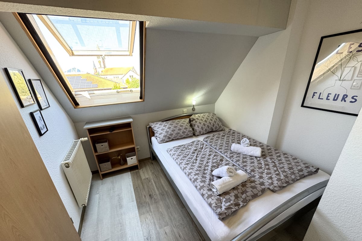A small, well-lit bedroom with a skylight, a bed with patterned bedding, a bedside table, a radiator, and a wall-mounted shelf.