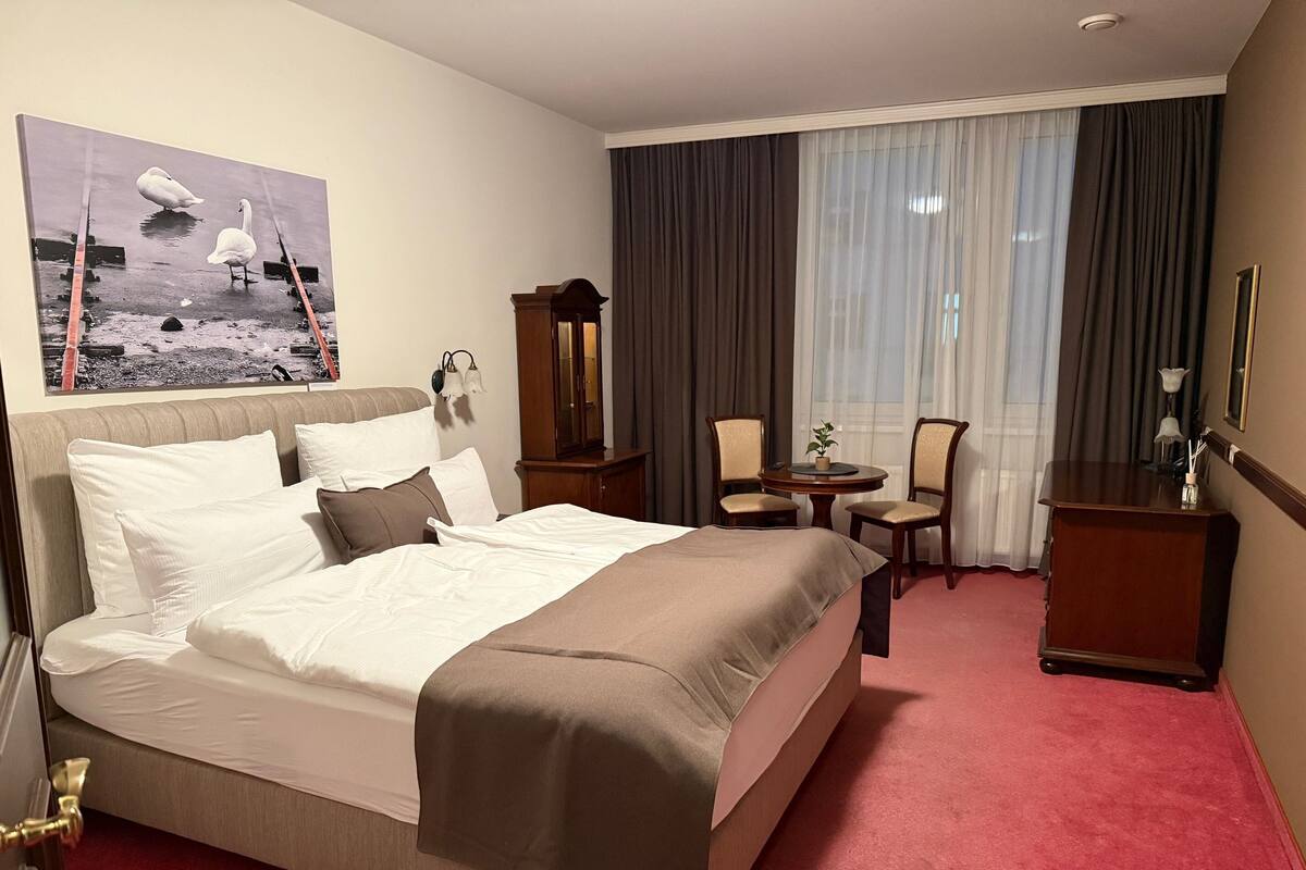 A hotel room with a large bed, a desk, two chairs, a small table, a mirror, and a painting of swans on the wall.