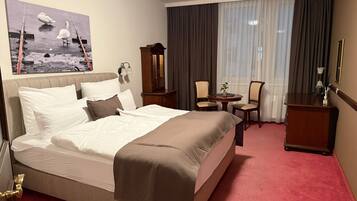 A hotel room with a large bed, a desk, two chairs, a small table, a mirror, and a painting of swans on the wall.