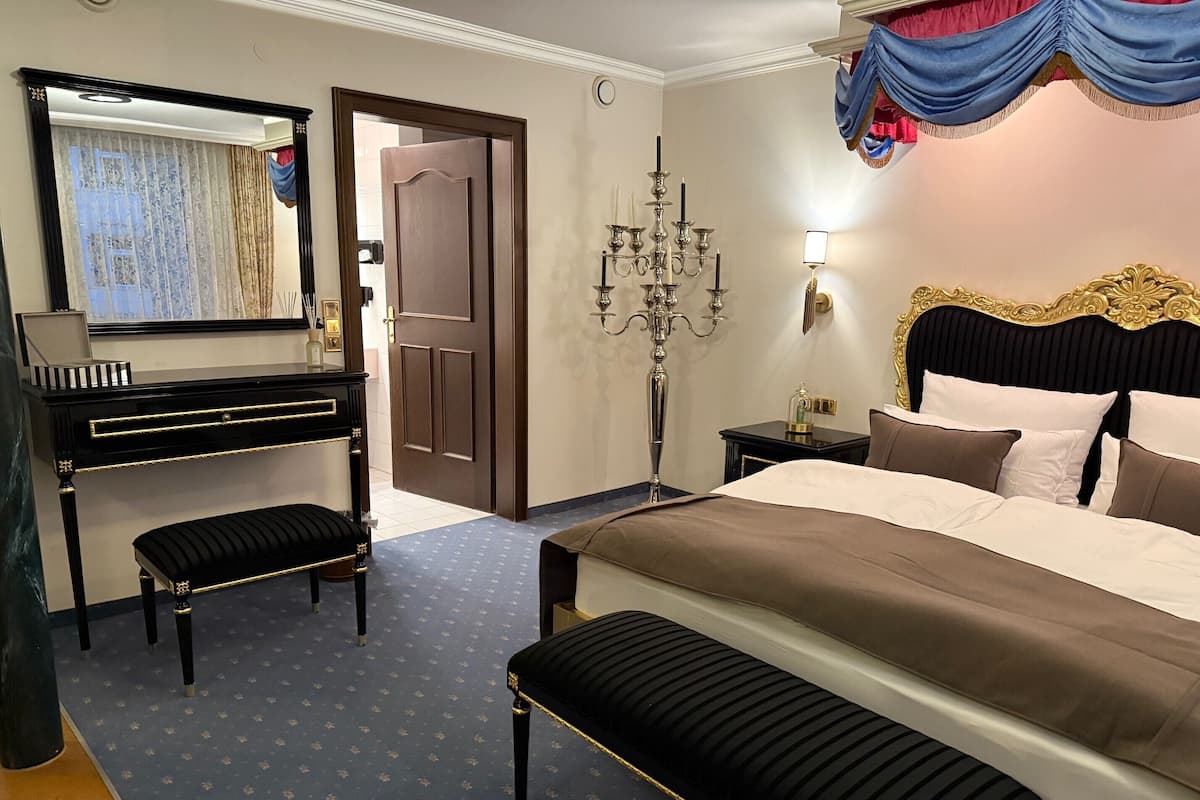 A hotel room with a large bed, a dresser, a bench, a mirror, a chandelier, and a door.