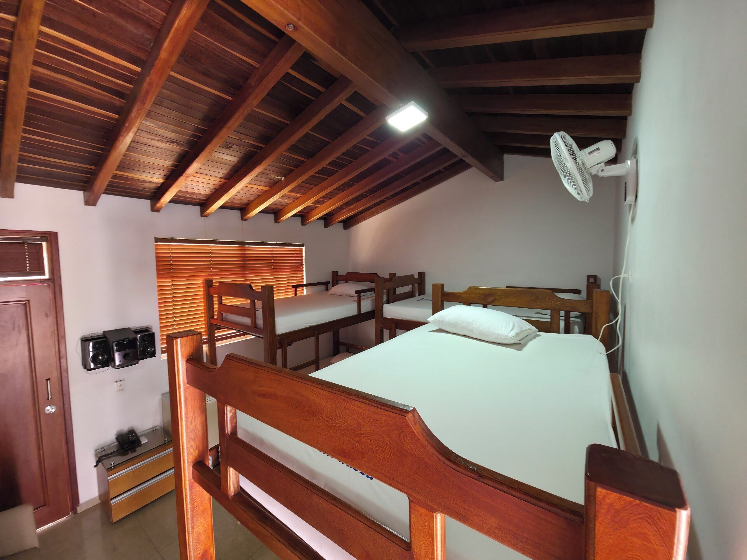 Quarto com beliche, vigas de madeira no teto, ventoinha e janela com persianas.