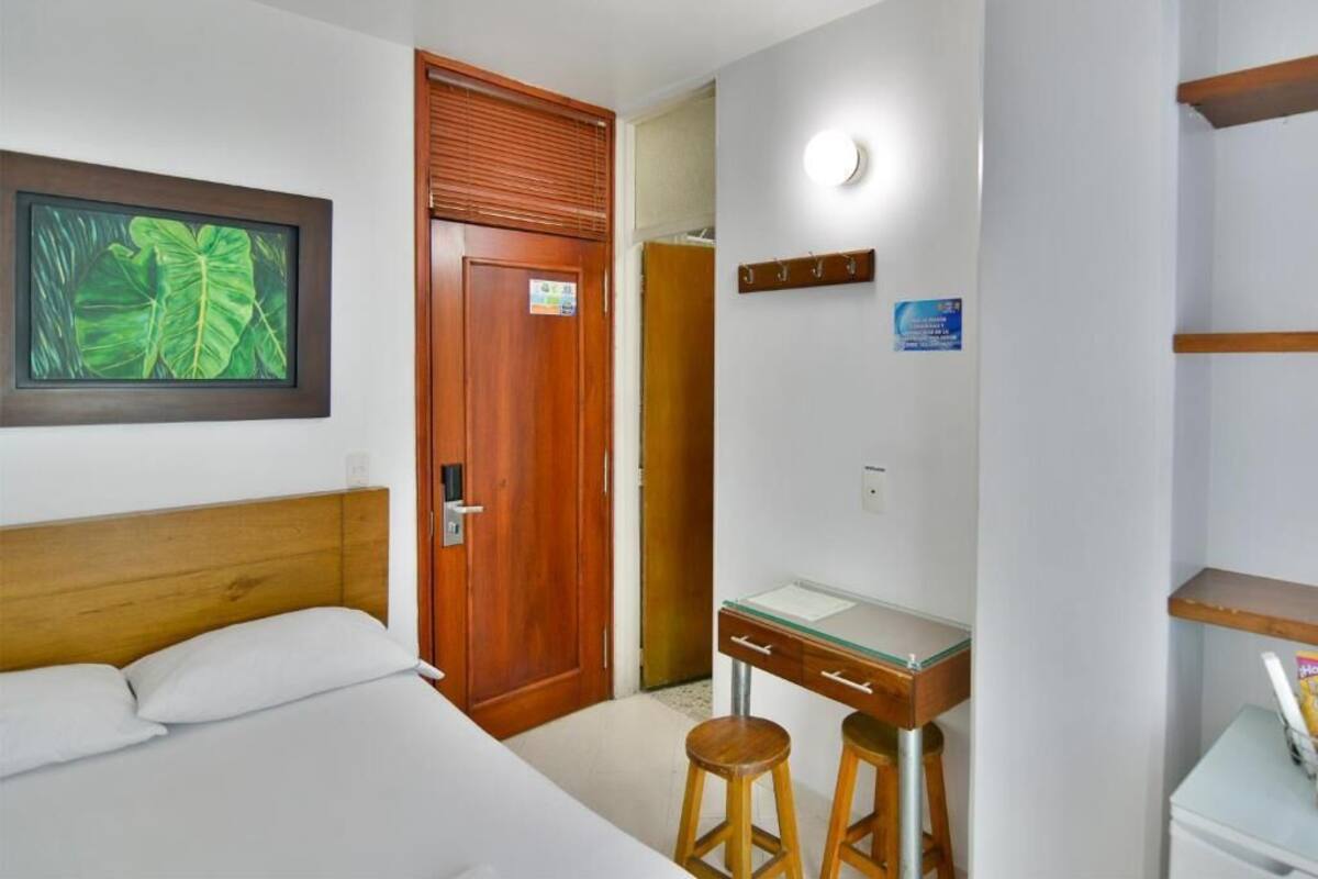 A hotel room with a bed, a wooden door, a desk with a chair, and a small stool.