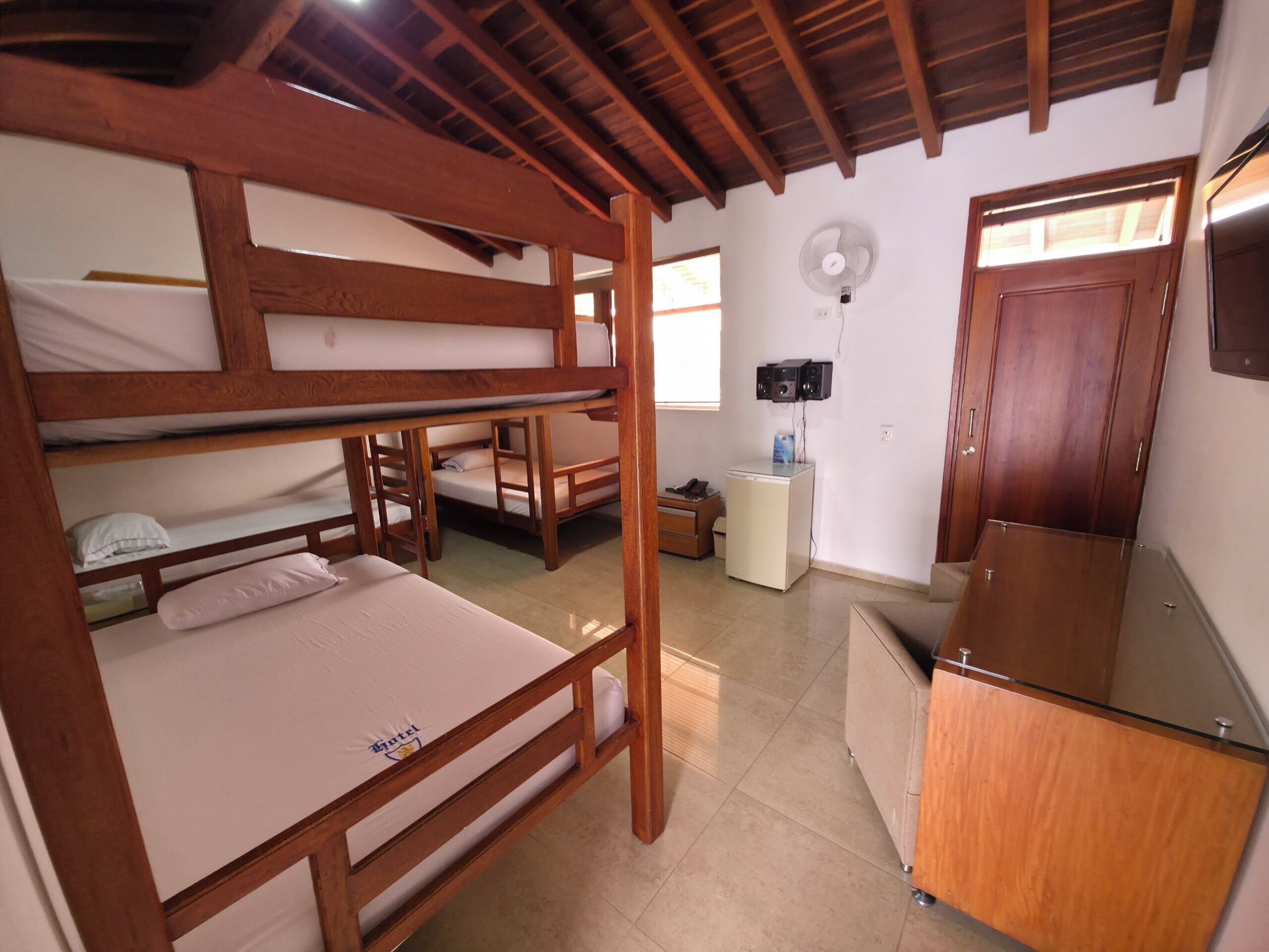 Une chambre avec des lits superposés, un petit réfrigérateur, un ventilateur et une porte en bois.