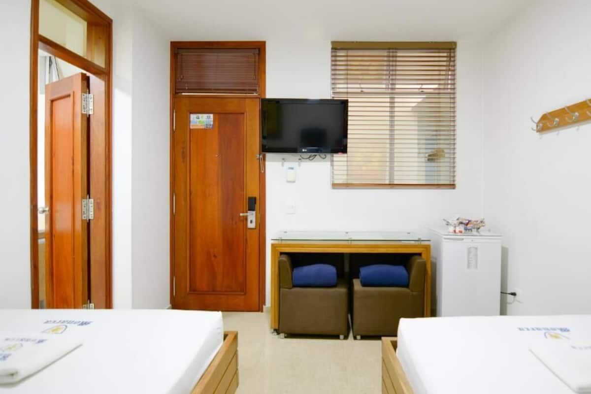 A room with two beds, a television, a desk with a computer, and a window with blinds.