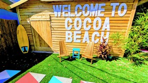 A wooden sign reading "WELCOME TO COCOA BEACH" with two lounge chairs and a surfboard in front. - King Studio Steps from Sand + Resort Backyard (Cocoa Beach)