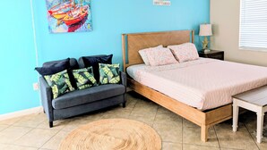 A bedroom with a bed, a sofa, a painting, and a ceiling fan. - King Studio Steps from Sand + Resort Backyard (Cocoa Beach)