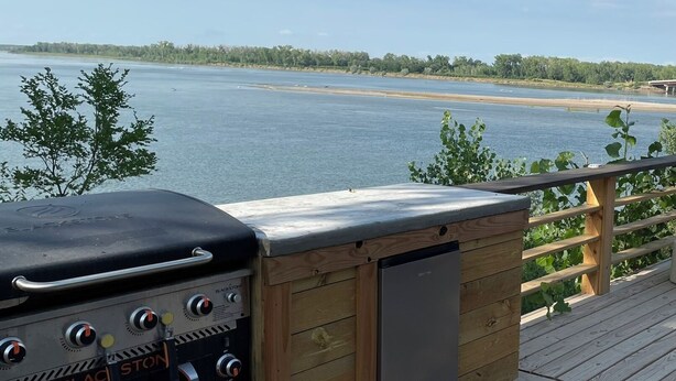 A wooden deck with a grill and a refrigerator, overlooking a calm lake. - Cozy RV with Expansive River Views (Newcastle)