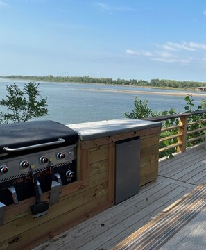 Una terrassa de fusta amb una barbacoa i un frigorífic, amb vista a un llac tranquil.