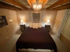 A bedroom with a large bed, two bedside tables with lamps, a ceiling light fixture, and a window with curtains.