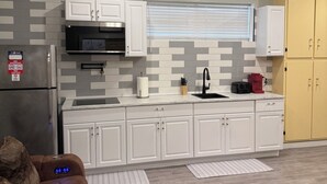 A compact kitchen with white cabinets, a black microwave, and a built-in oven. There is a window with blinds, a sink, and a countertop with various items.
