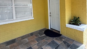 A covered porch with yellow brick walls, a white door, and a window with blinds.