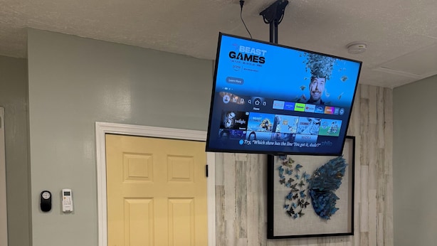 A hotel room with a bed, a desk with two stools, a mounted TV displaying a game streaming service, and a door.