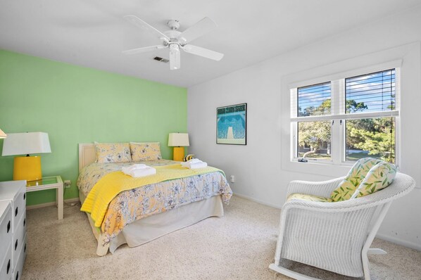 A bedroom with a bed, two bedside lamps, a ceiling fan, a window with blinds, and a chair. - Oceanfront, Pet Friendly, Golf Cart included - 711 Bluefish (Fripp Island)