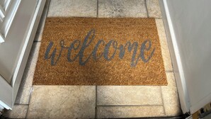A welcome mat with the word "welcome" in cursive on a brown doormat at the entrance of a hallway with tiled flooring and white walls. - 4-6 Guests in centre location with stunning views (Llanberis)