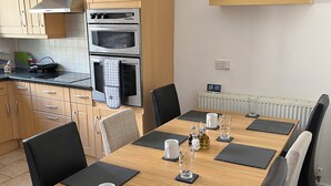 A dining area with a wooden table set for six, surrounded by black chairs. A kitchen with wooden cabinets and stainless steel appliances is visible in the background. - 4-6 Guests in centre location with stunning views (Llanberis)