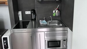 A compact kitchenette with a stainless steel stove, oven, and microwave, a sink with a faucet, and a small shelf with a coffee maker and a vase of flowers. - Tiny House "Black Pearl" (Tettnang)