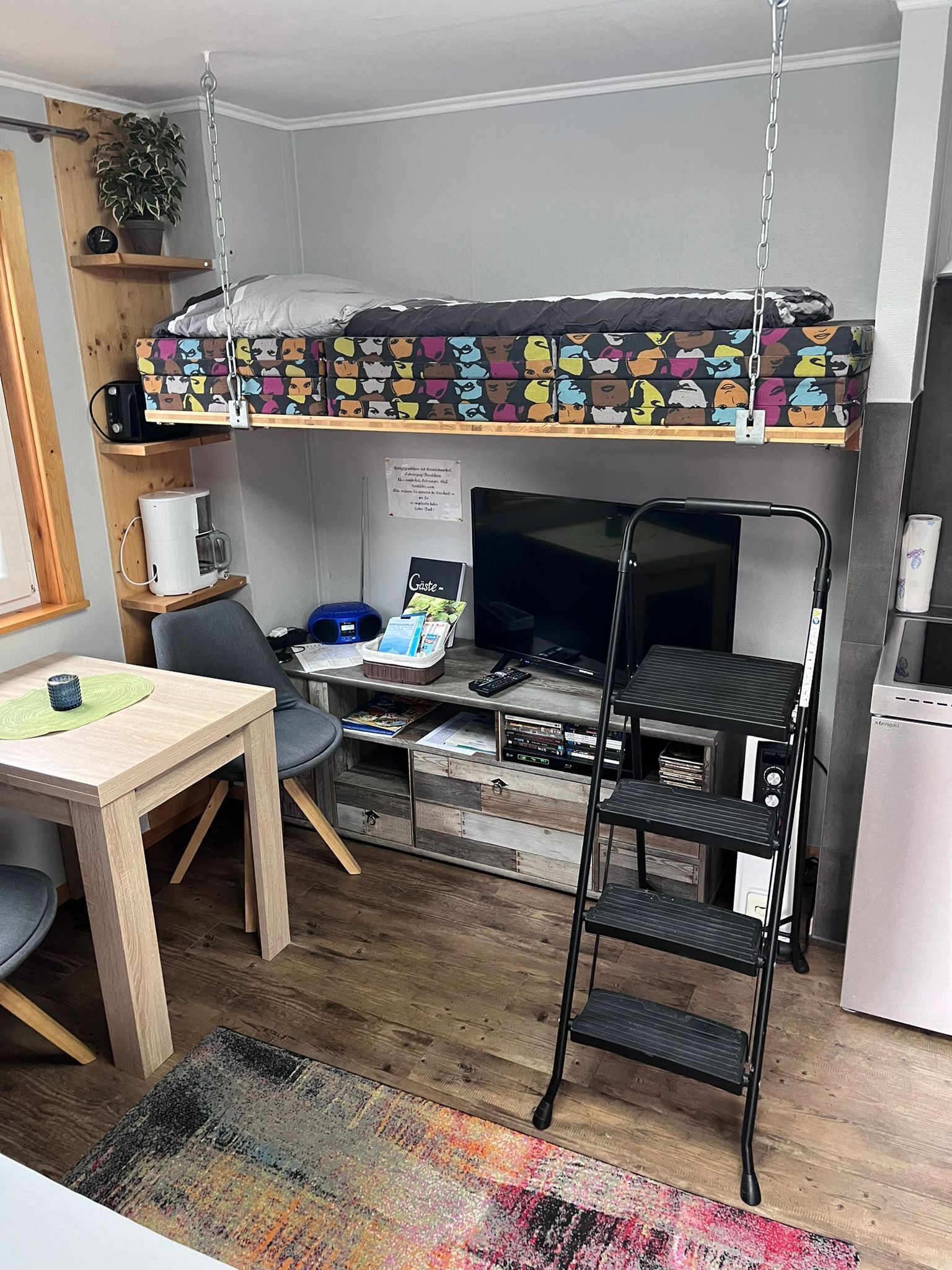 Een kleine kamer met een stapelbed, een bureau met stoel, een koffieapparaat, een televisie en een kitchenette.