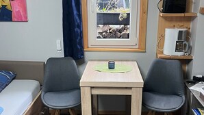 A small, cozy room with a bed, two chairs, a table, a window with blinds, a shelf with a coffee maker, and a painting on the wall. - Tiny House "Black Pearl" (Tettnang)