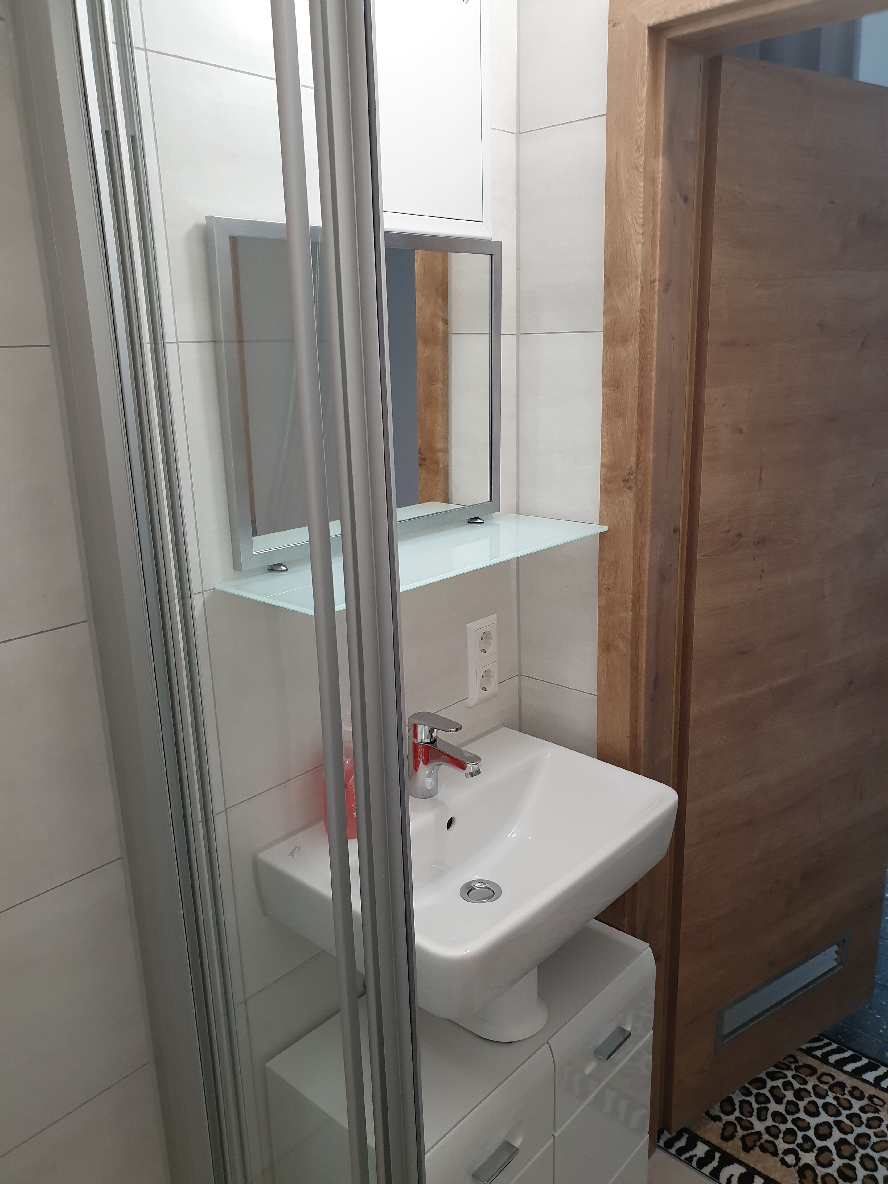 A modern bathroom with a glass shower enclosure, a white sink, and a wooden door.