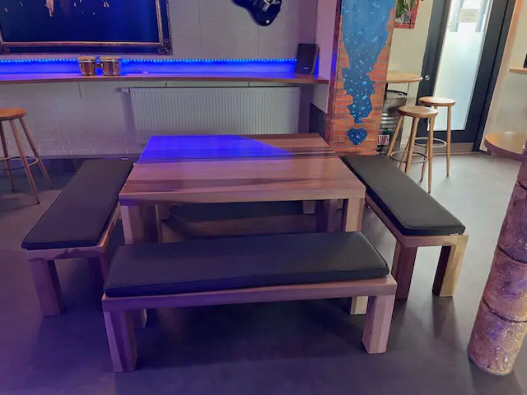 Eine hölzerne Bank mit gepolstertem Sitz und Rückenlehne, einem kleinen Tisch und einem an der Wand befestigten Lautsprecher.
