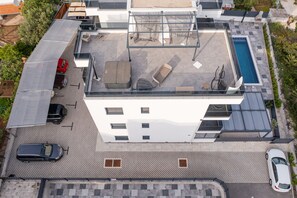 An aerial view of a modern building with a rooftop pool, multiple cars parked, and a covered outdoor seating area.
