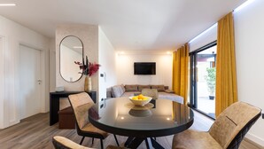 Una sala de estar moderna con una mesa redonda de vidrio, cuatro sillas, un sofá, un televisor de pantalla plana y un ventanal con cortinas amarillas.
