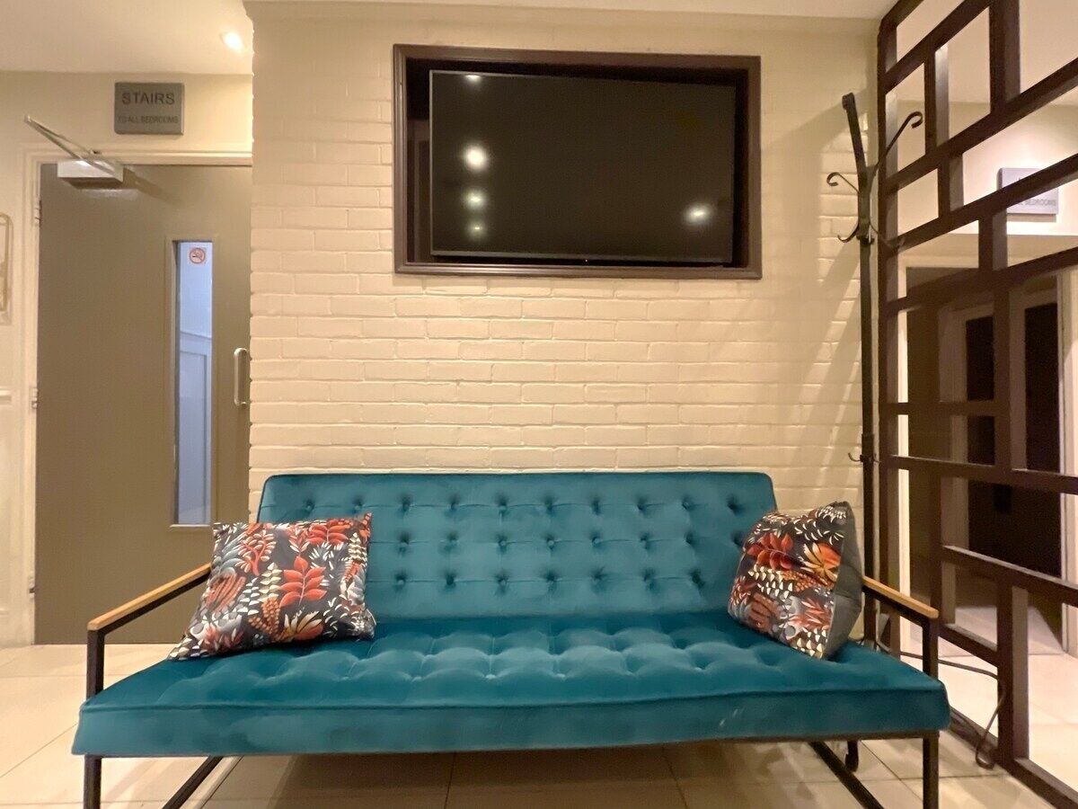A teal sofa with floral cushions in a room with a white brick wall and a large flat-screen TV mounted above it.