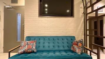 A teal sofa with floral cushions in a room with a white brick wall and a large flat-screen TV mounted above it.