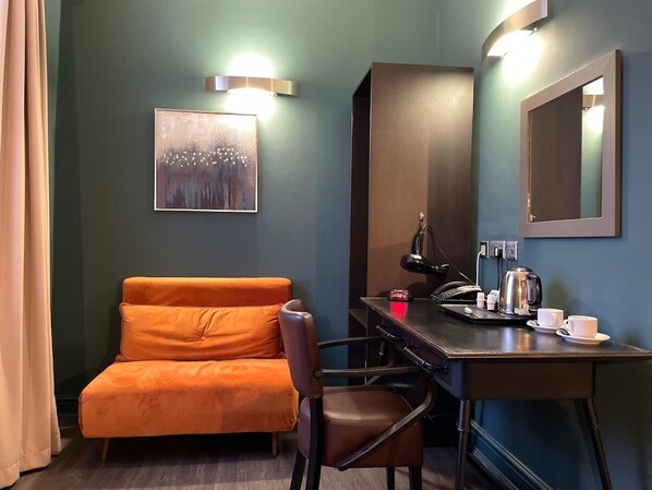 A hotel room with a sofa, a desk with a lamp, a coffee maker, and a framed picture on the wall.
