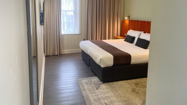 Una habitación de hotel moderna con una cama grande, un ventanal con cortinas, una mesita de noche con lámpara y piso alfombrado.