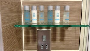 A bathroom with a glass shelf holding six bottles of hand and body lotion, a shaver-only outlet, and a towel warmer. - HNFC Stays - Business En-Suit (Tyne and Wear)