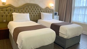 A hotel room with two beds, a large tufted headboard, a bedside table with a lamp, and a window with curtains.