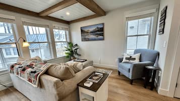 A cozy living room with a sofa, armchair, and coffee table, large windows with blinds, and a painting on the wall.