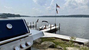 Un bote atracado en un muelle con una bandera estadounidense, una parrilla y un lago tranquilo al fondo.
