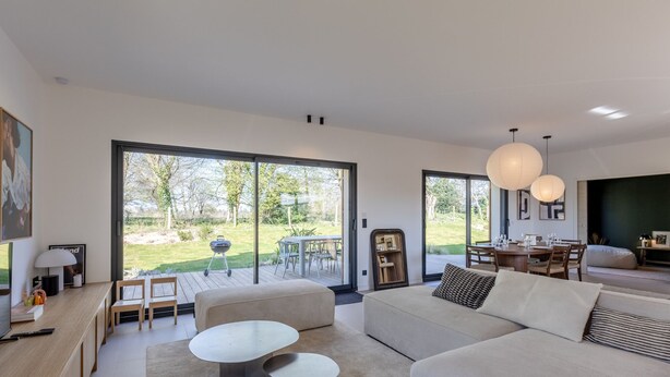 A modern living room with a large sliding glass door leading to a garden, a sofa, a dining table, and a TV. - L'Élégance | 115 m² | 4 + 2 guests | Design & Nature (Honfleur)