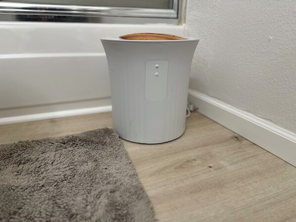Ein weißes, zylinderförmiges Objekt mit Bedienfeld auf Holzboden neben einer weißen Wand und einem Fenster.