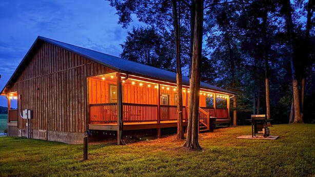 A rustic wooden cabin with a porch and string lights, surrounded by trees and grass. - Escape to the Farm- Minutes from Dale Hollow Lake! (Alpine)