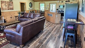A spacious living room with a brown leather sofa, wooden flooring, a ceiling fan, and a kitchen area with wooden cabinets and appliances. - Escape to the Farm- Minutes from Dale Hollow Lake! (Alpine)