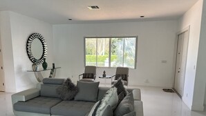 A modern living room with a grey sectional sofa, a glass coffee table, and a large mirror on the wall. There are two chairs and a small table with decorative items.