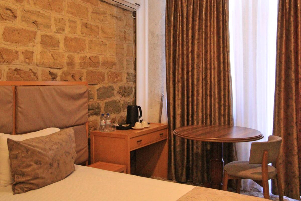 A hotel room with a stone wall, a bed with pillows, a wooden desk with a coffee maker, a table with a chair, and a window with curtains.