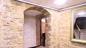 Un pasillo con paredes de piedra, suelo de baldosas, un espejo enmarco y una escalera con barandilla de madera.